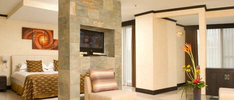 Quarto de hotel moderno com parede de detalhes em pedra, TV de tela plana, sofá confortável, mesa de centro de vidro e um vaso decorativo com flores.