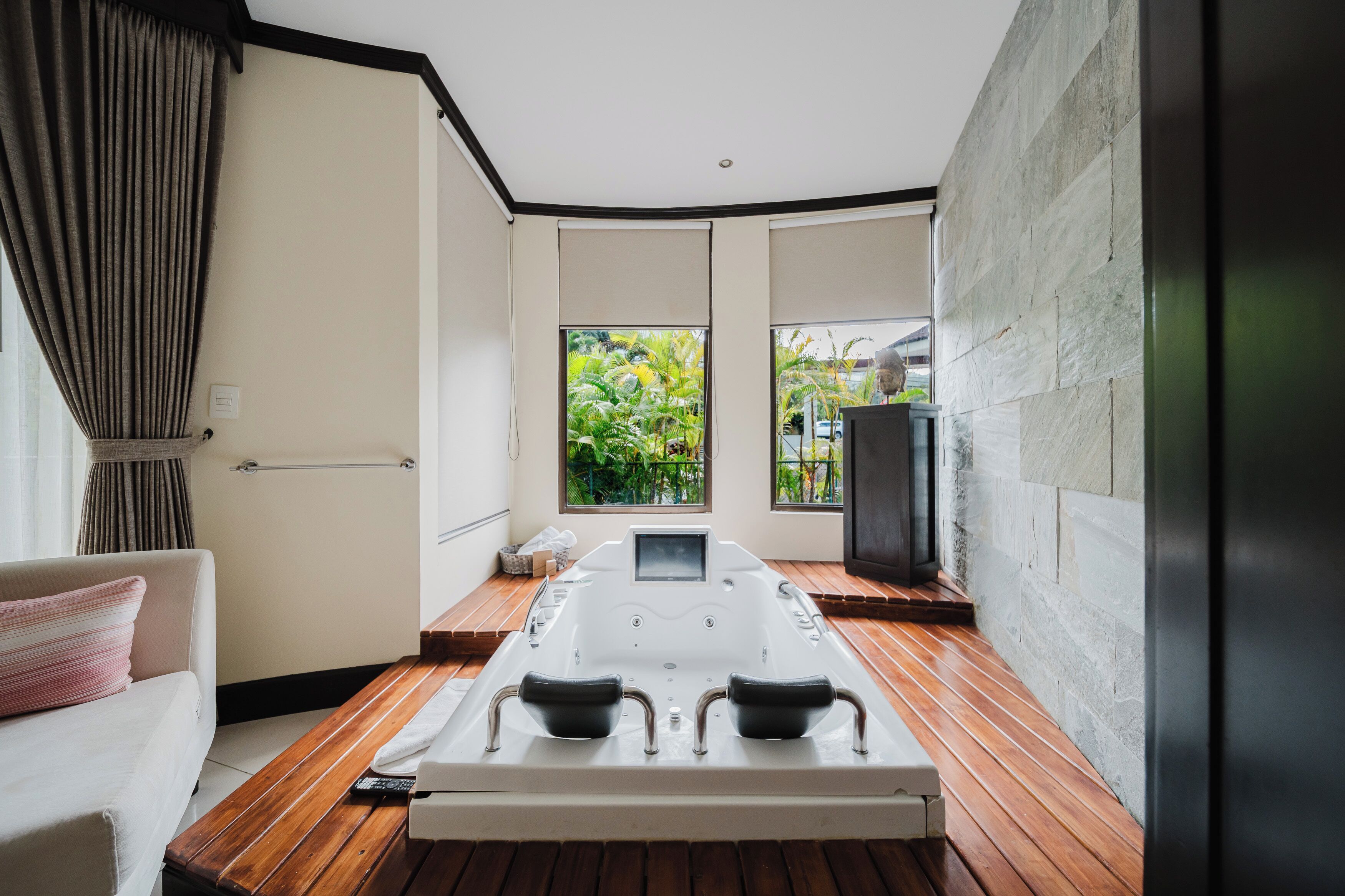 A modern spa room with a wooden floor, a massage table, a sofa, and large windows offering a view of greenery.