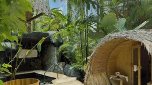 A thatched-roof hut with a small porch, a stone wall, and a wooden tub in a tropical garden setting.