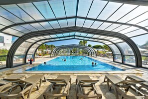 An indoor swimming pool with clear glass ceilings, lounge chairs, and a view of the surrounding landscape.