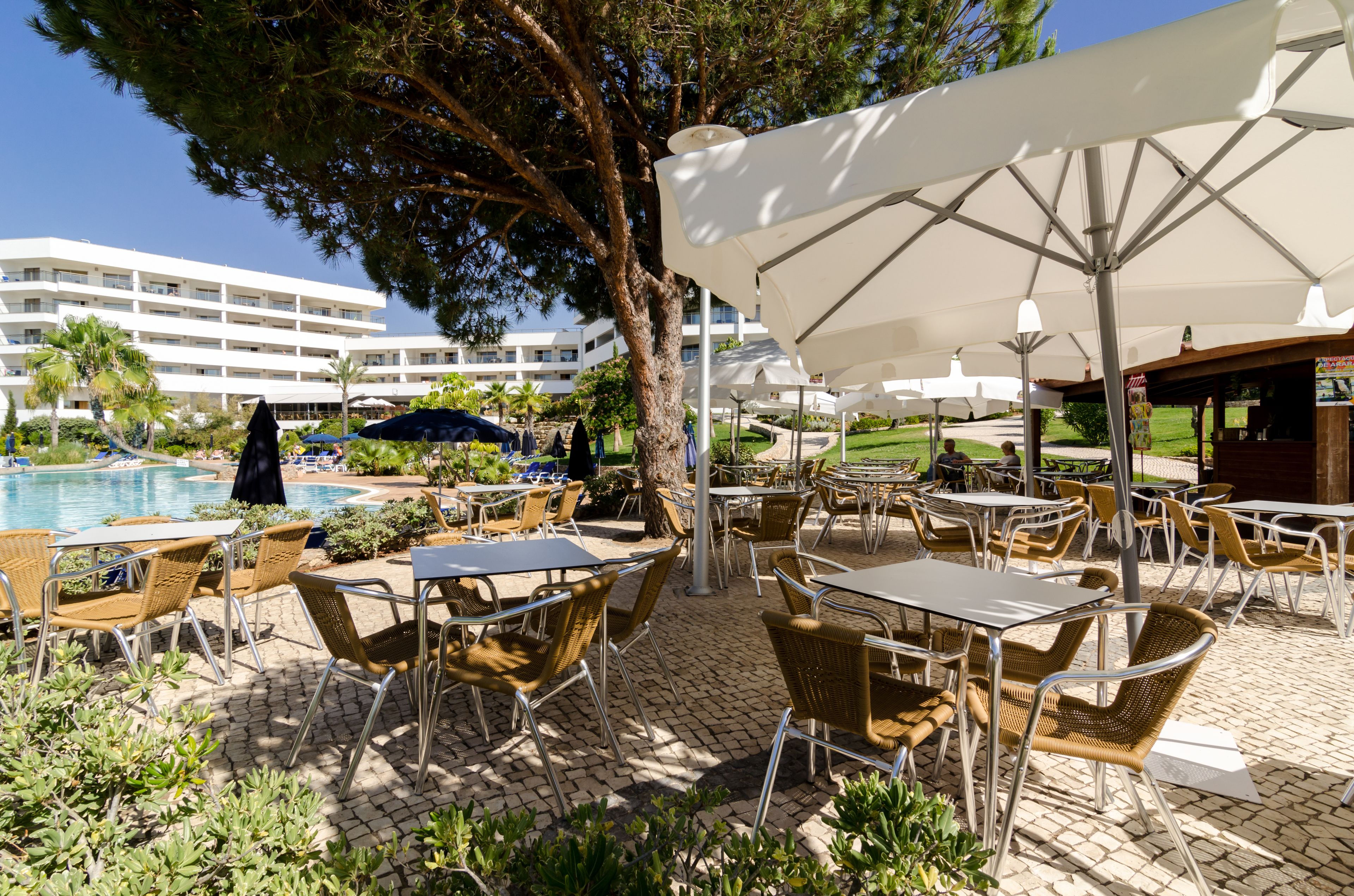 Een terras buiten met tafels, stoelen, parasols en een zwembad op de achtergrond.