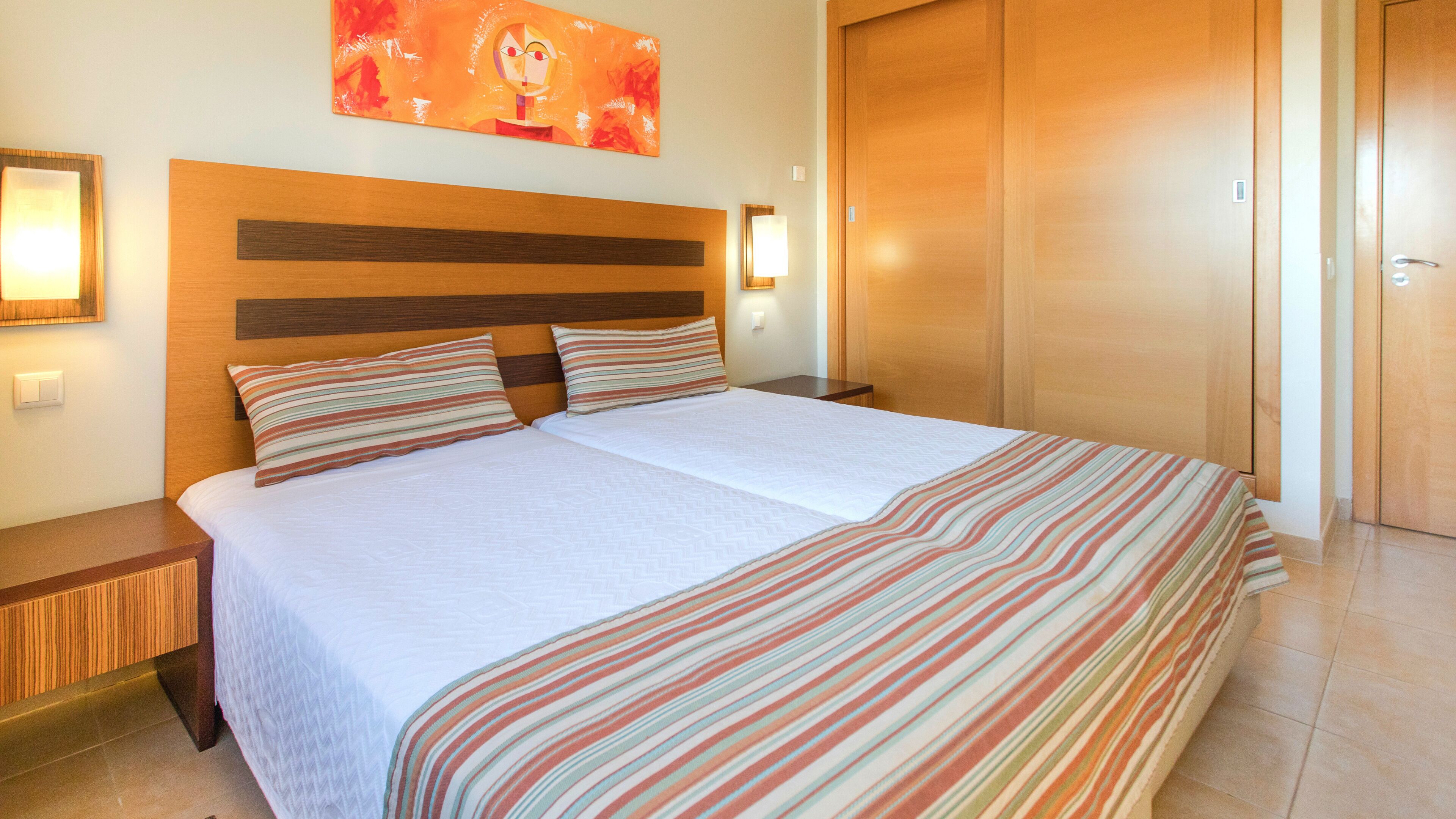A double bed with striped bedding, a wooden headboard, a nightstand with a lamp, and a painting on the wall.