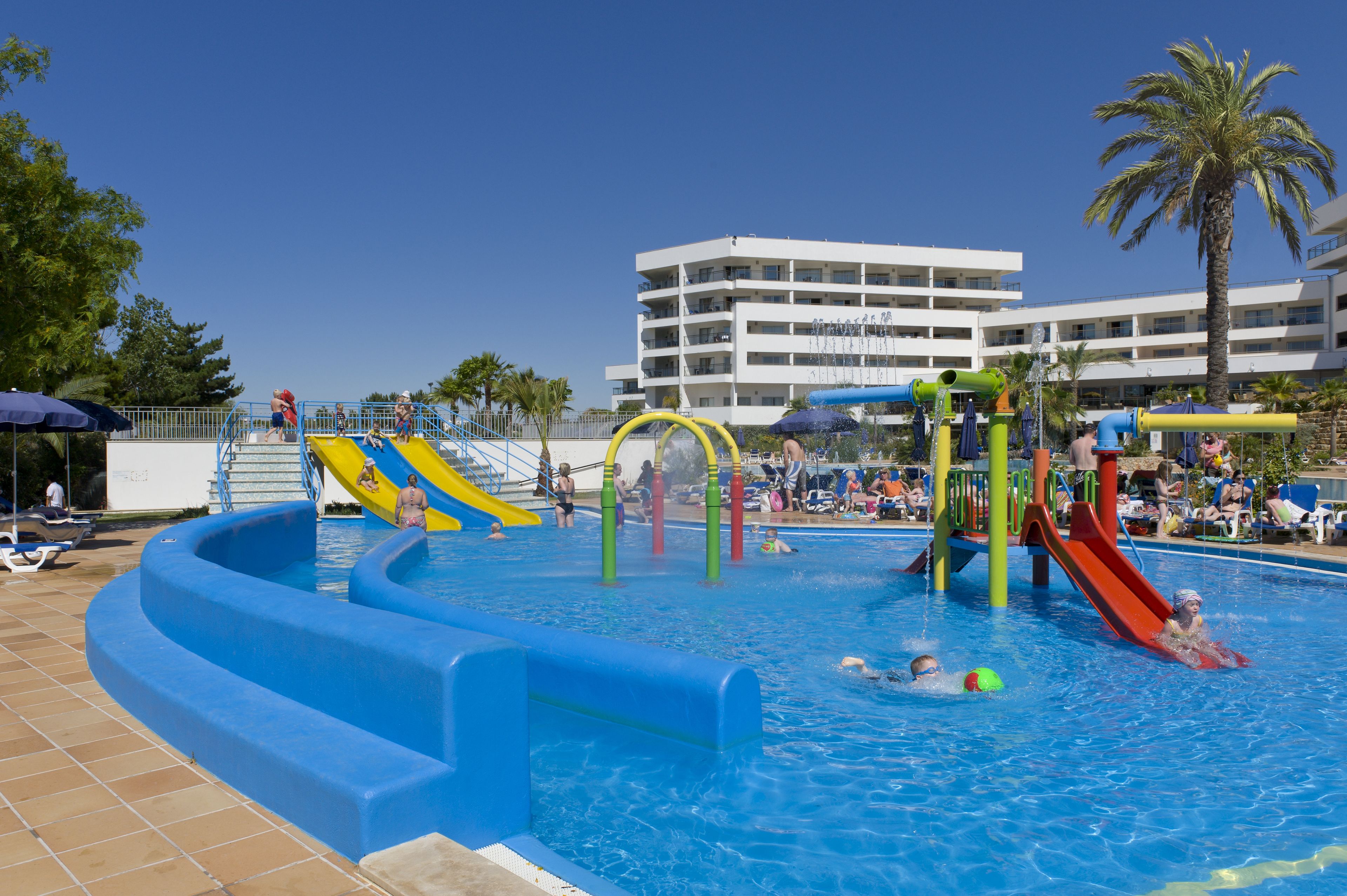 A swimming pool with slides and water play structures, surrounded by a tiled deck and palm trees.