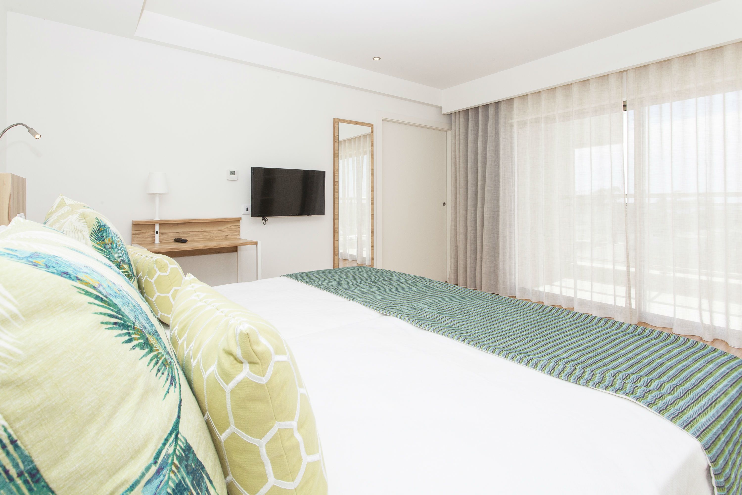 Een hotelkamer met een groot bed, een bureau met een flatscreen televisie en een raam met dunne gordijnen.