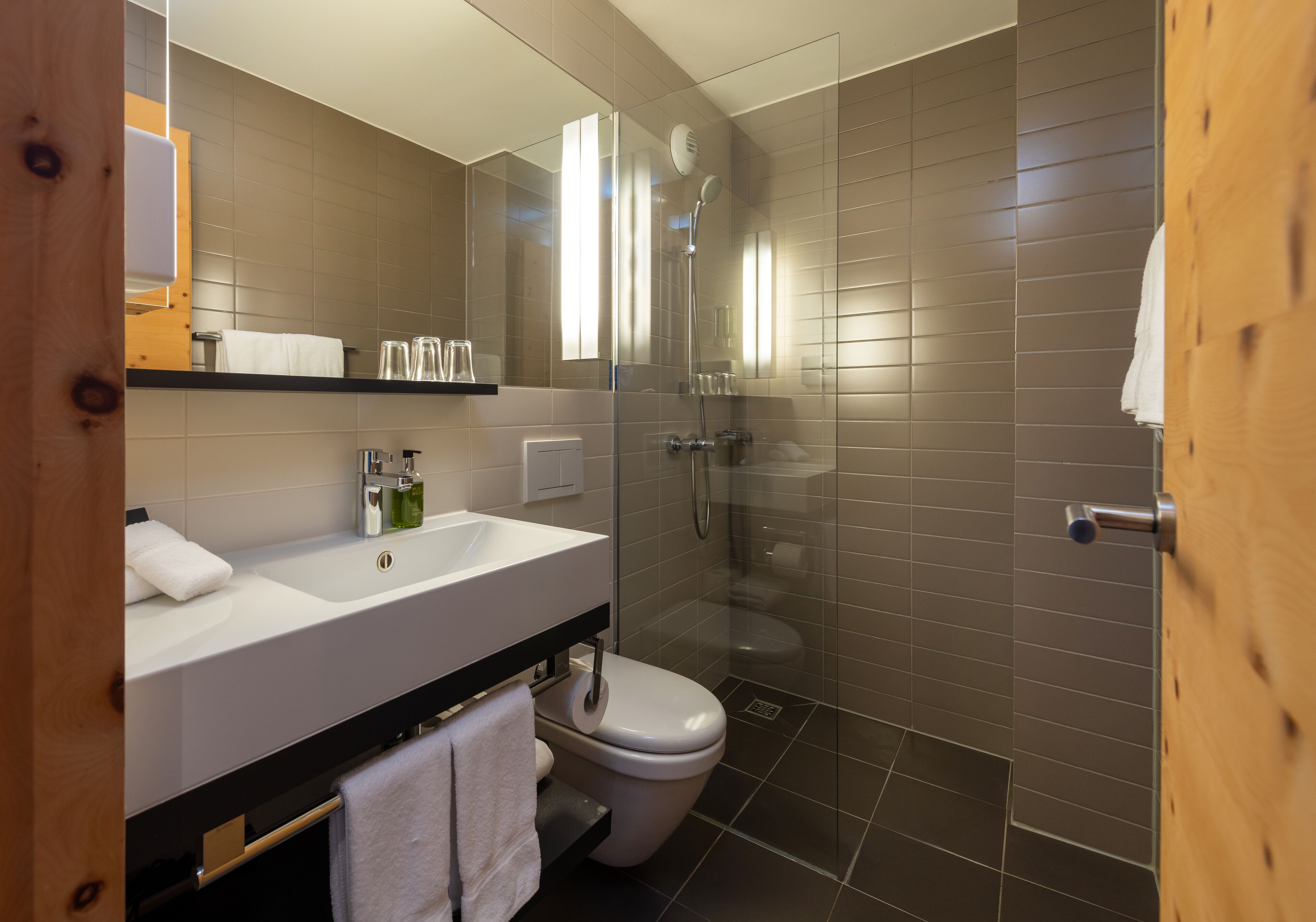 A modern bathroom with a white sink, mirror, and toilet. It includes a glass-enclosed shower and a wooden door.
