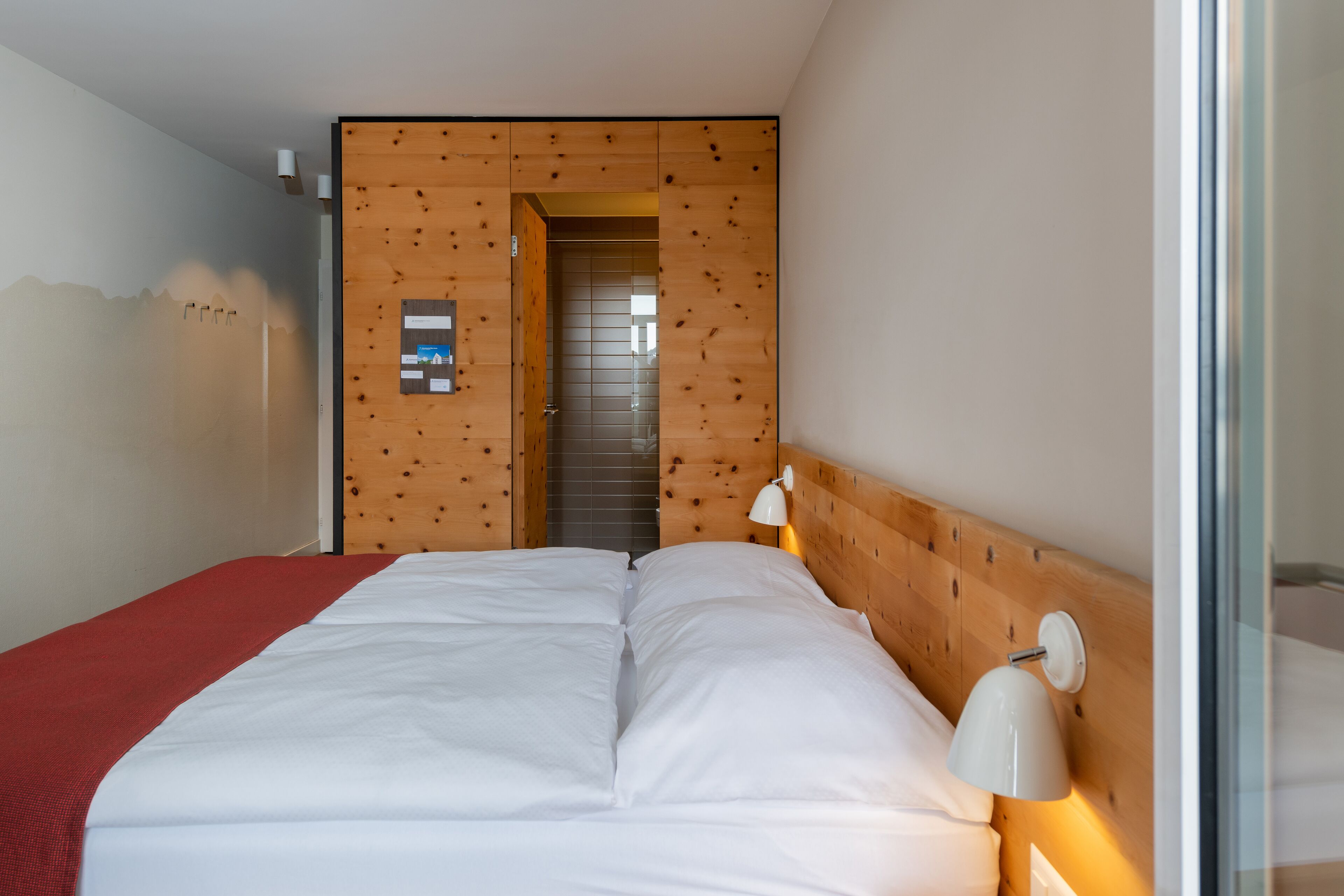 A modern hotel room with a wooden headboard, a large bed with white linens, and a small bedside table with a lamp.