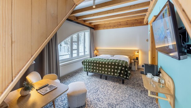 A modern hotel room with a wooden ceiling, a bed with a green patterned bedspread, a desk with a remote control, a small potted plant, and a view of the outdoors through a large window. - Schweizerhof Sils Maria, a Faern Collection Hotel (Sils im Engadin-Segl)
