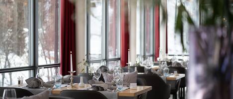 Um restaurante com mesas postas para refeições, grandes janelas e cortinas vermelhas.