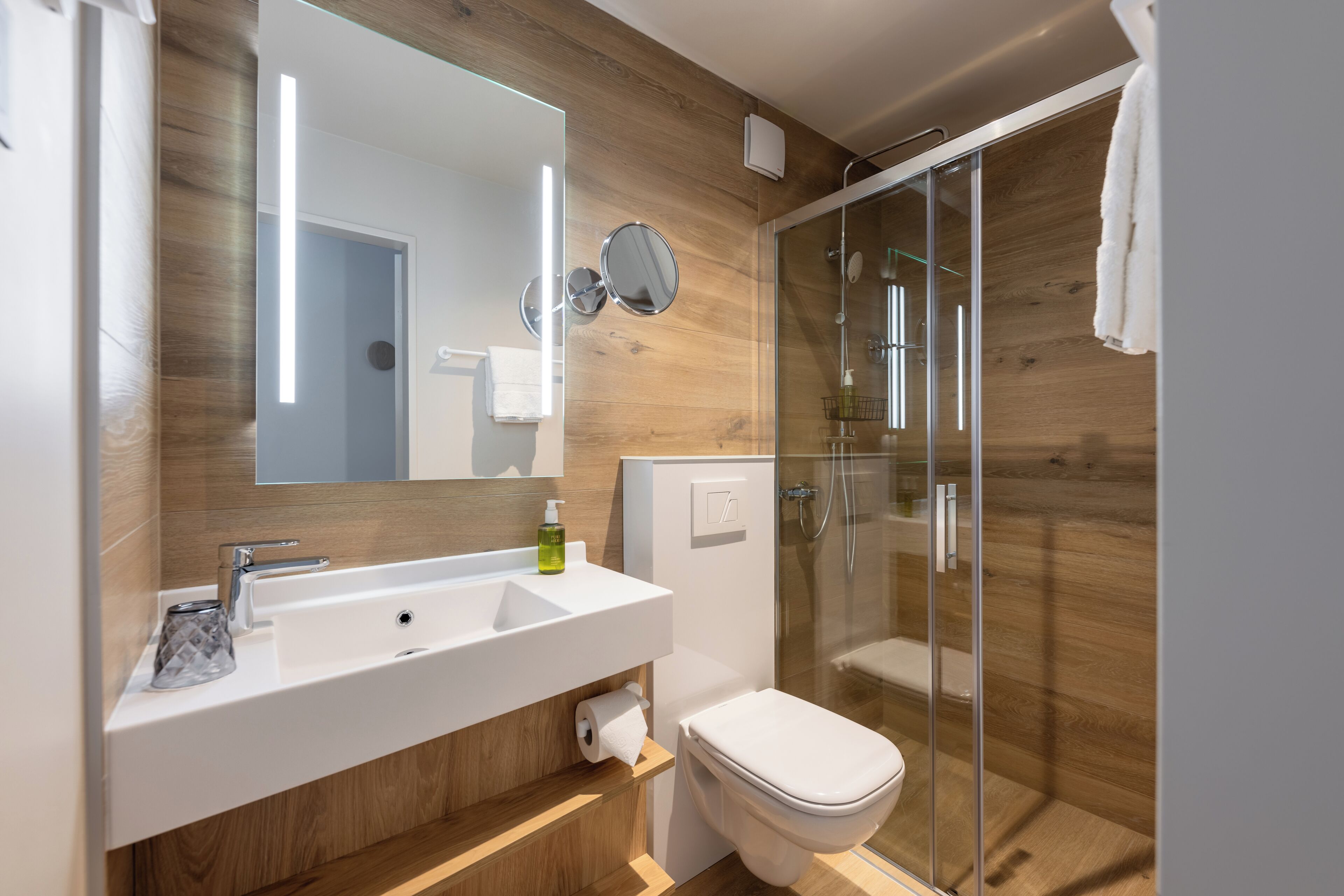 Un baño moderno con pared de madera, cabina de ducha de vidrio, lavamanos blanco y retrete.