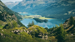 Paisaje montañoso con un lago, colinas boscosas y un pequeño asentamiento.