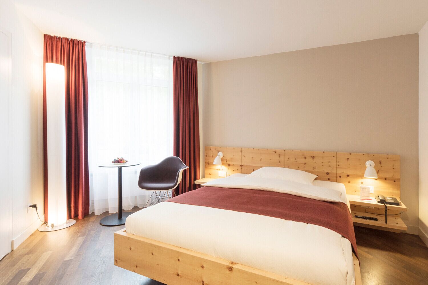 Une chambre d’hôtel avec un lit en bois, deux lampes de chevet, une petite table avec une chaise et une fenêtre avec des rideaux.