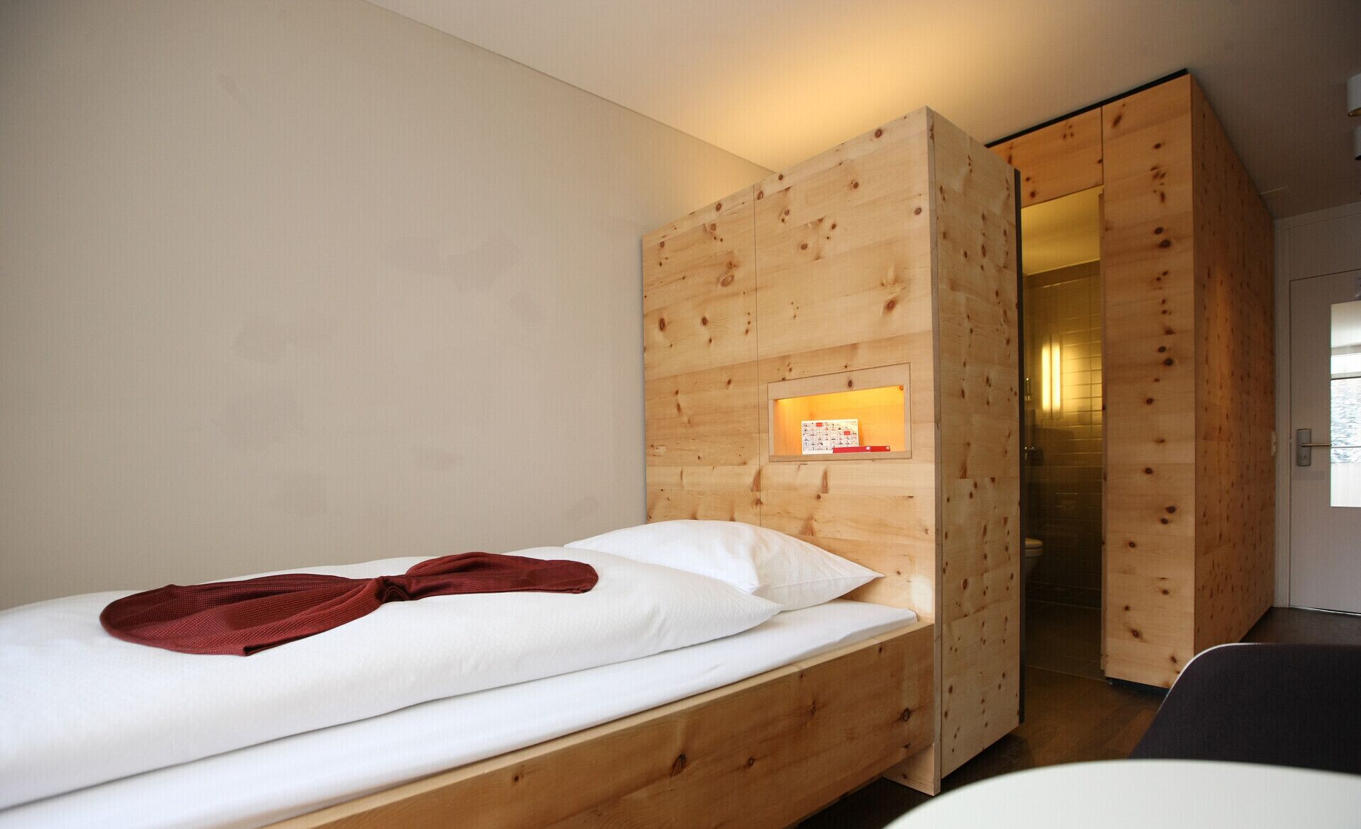 A single bed with a red blanket, wooden walls, and a small illuminated panel on the wall.