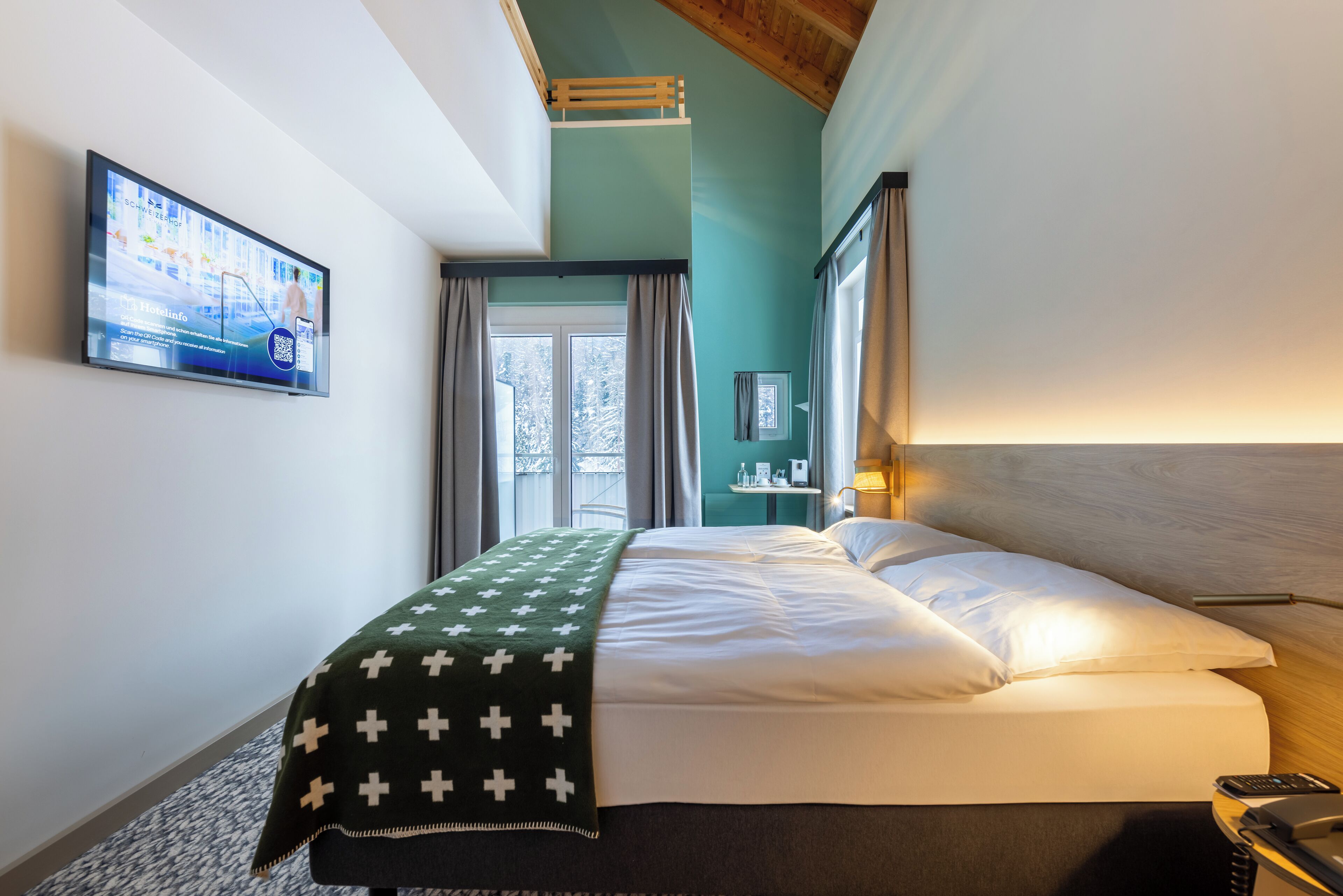 Ein Hotelzimmer mit einem Bett, einem an der Wand montierten Fernseher, einem Fenster mit Vorhängen und einer Holzbalkendecke.