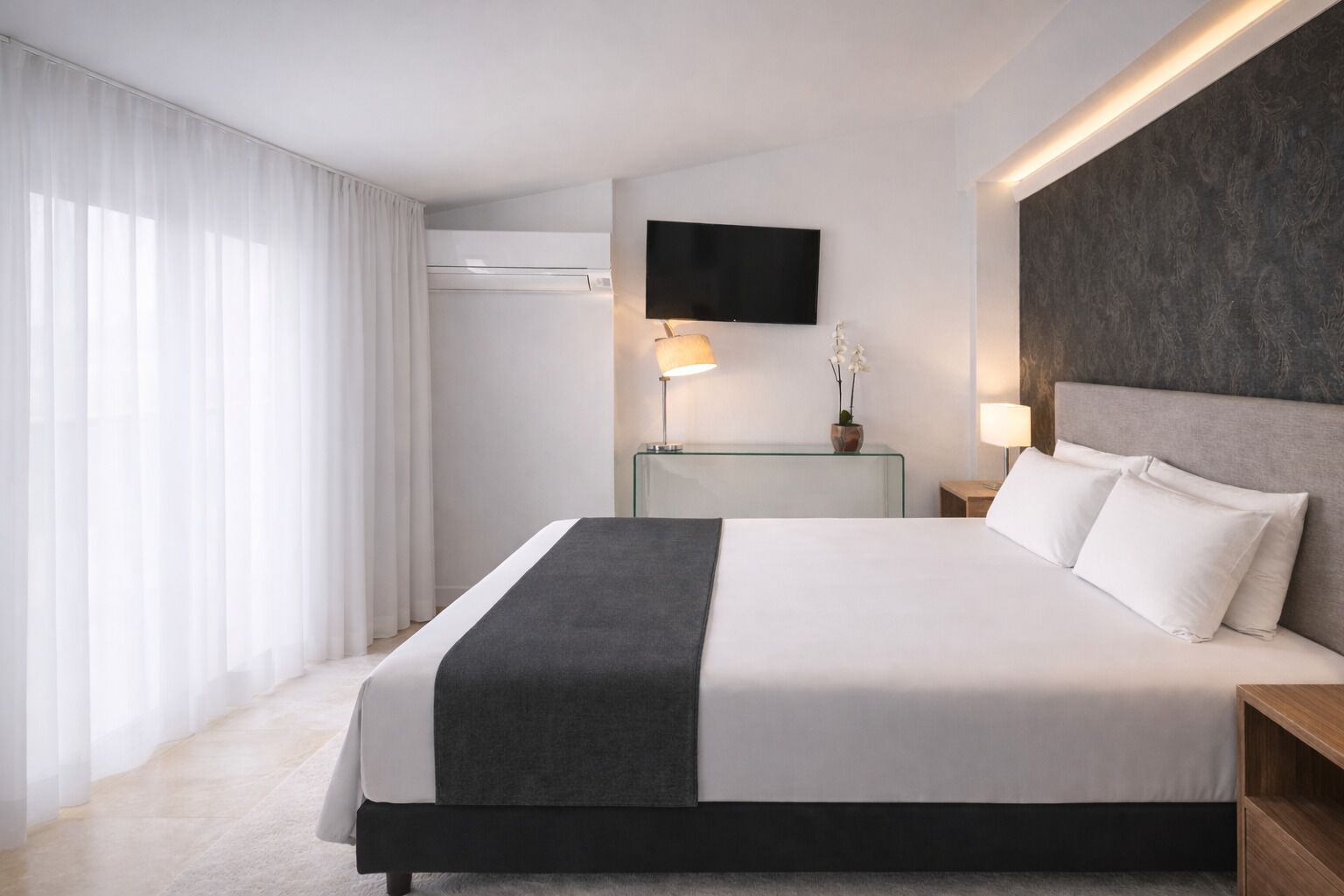 A modern hotel room with a large bed, a flat-screen TV mounted on the wall, a glass side table with a lamp, and a view of the outdoors through sheer curtains.