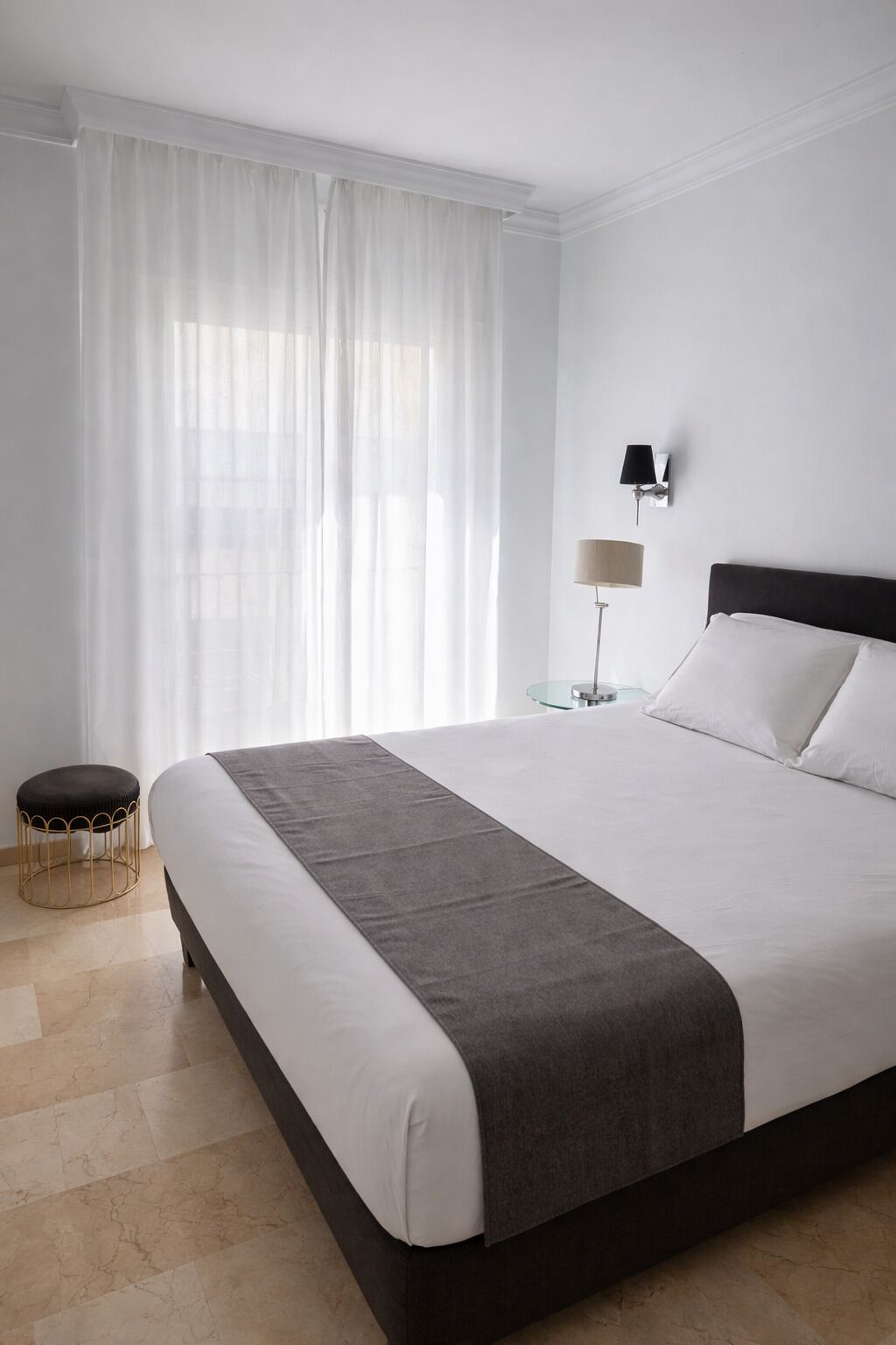 A modern hotel room with a large bed, a small round table, a floor lamp, and sheer curtains.