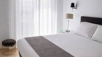 A modern hotel room with a large bed, a small round table, a floor lamp, and sheer curtains.