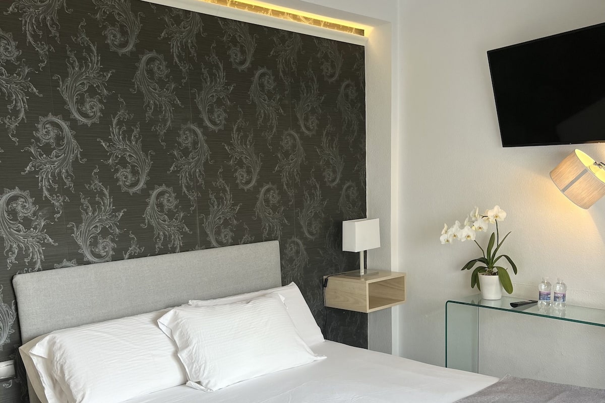 Una habitación de hotel moderna con una cama grande, un televisor de pantalla plana montado en la pared, una mesita de noche con lámpara y un pequeño florero con flores.