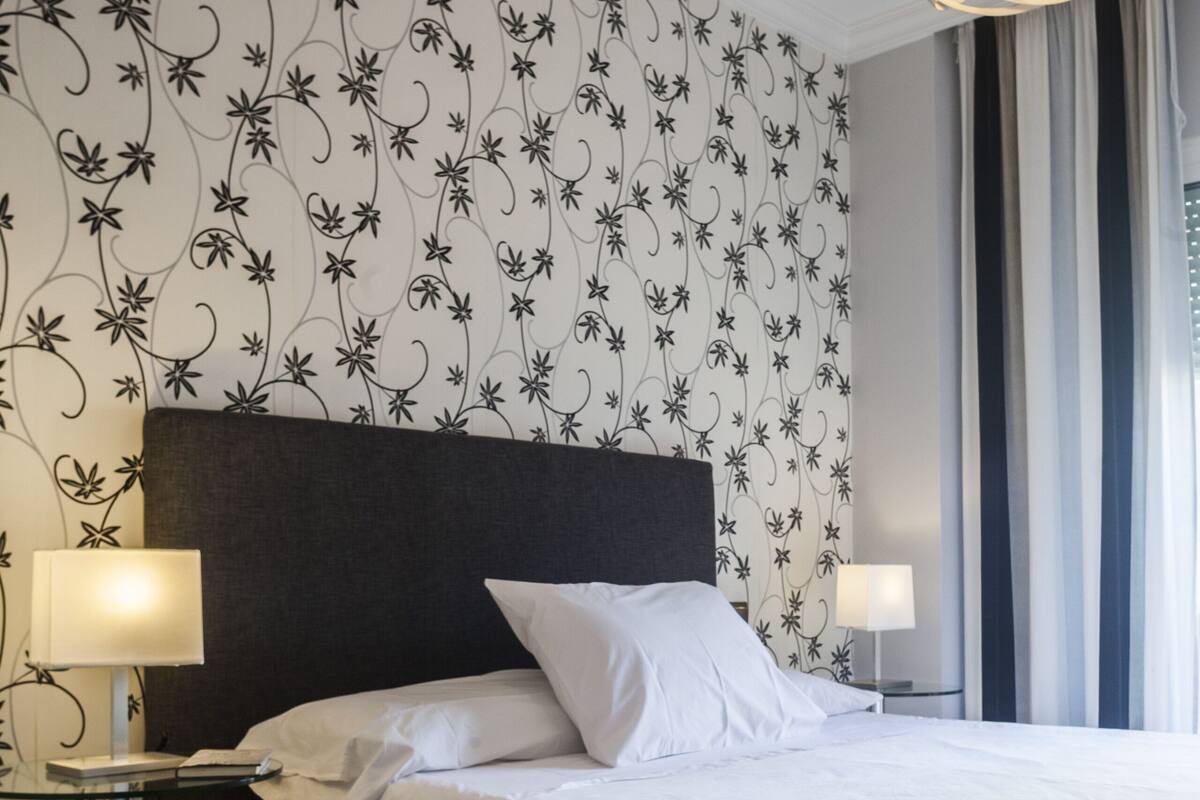 Habitación de hotel con cama, dos lámparas de noche, cabecera oscura, papel tapiz con motivos florales y una ventana grande con cortinas.