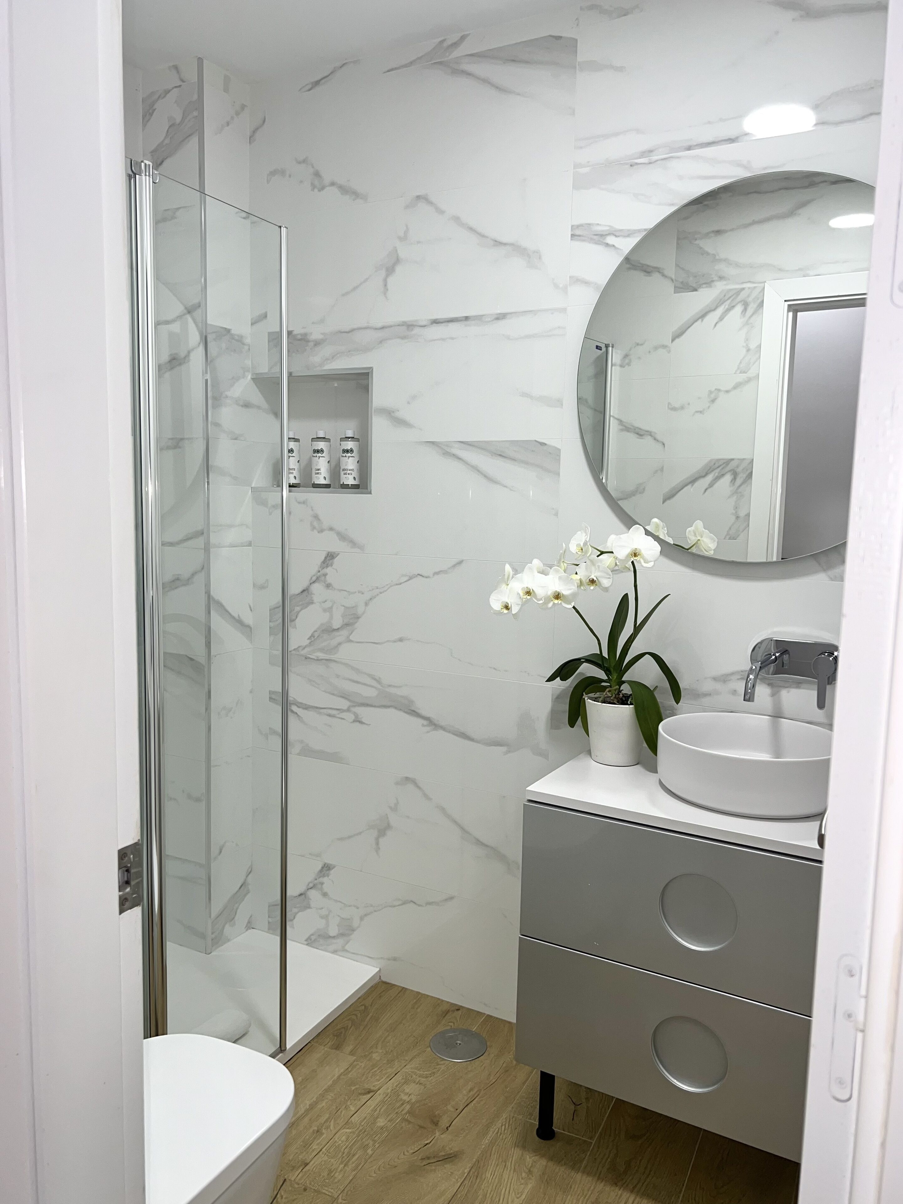 Une salle de bain moderne avec une douche à parois de verre, un mur recouvert de carreaux de marbre, un miroir rond, un lavabo agrémenté d’une orchidée en pot et un meuble-lavabo gris doté de tiroirs.
