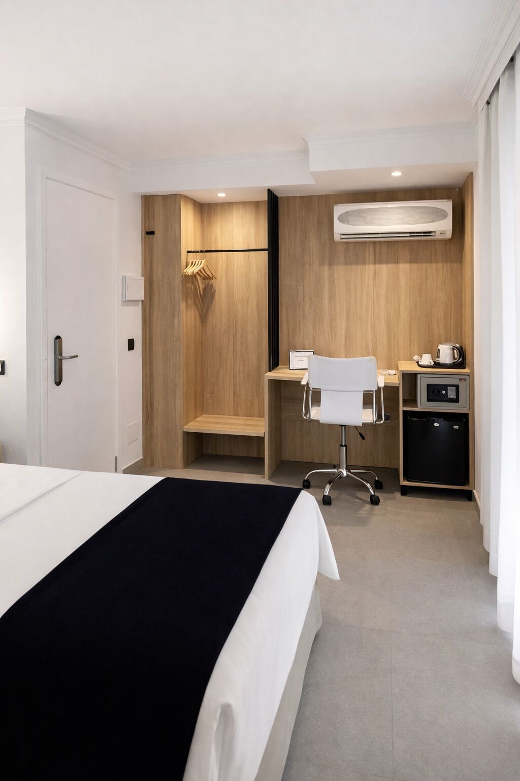 Habitación de hotel con cama, escritorio, silla, equipo de aire acondicionado y refrigerador pequeño.