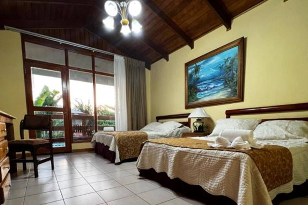 A room with two beds, a wooden ceiling, and a large window with a view of greenery.