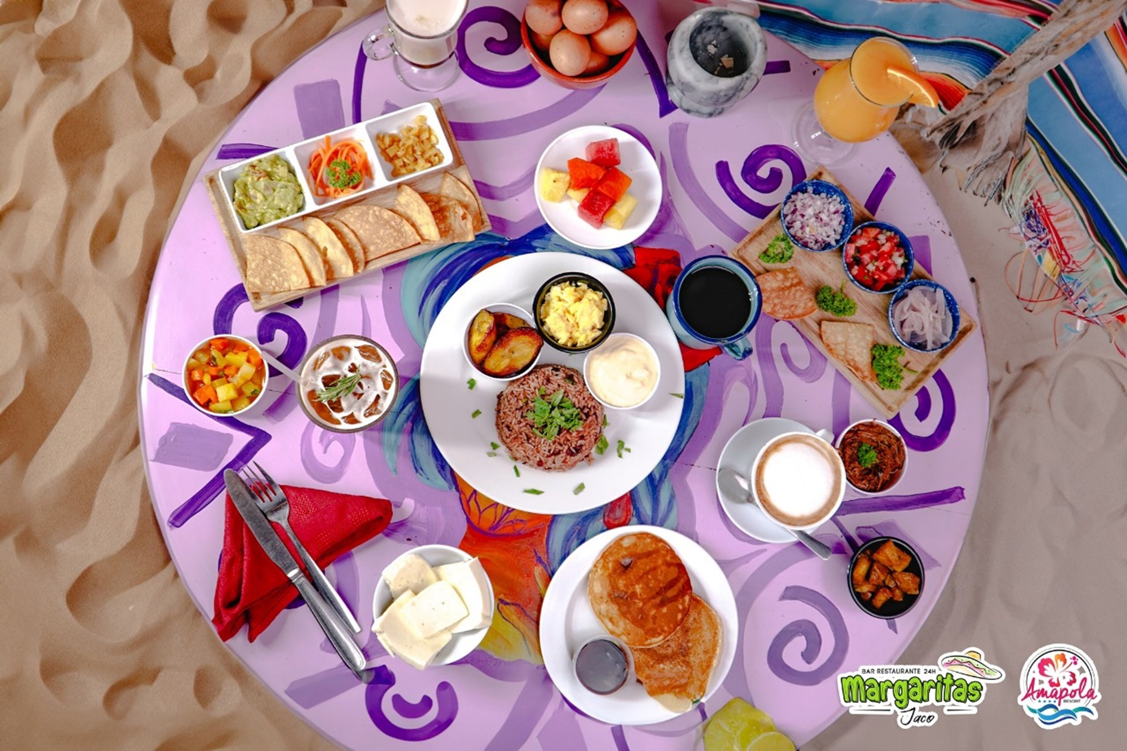 Une table colorée, dressée avec une nappe à motifs, où l’on trouve une variété de plats, notamment des tacos, du riz et différentes boissons.