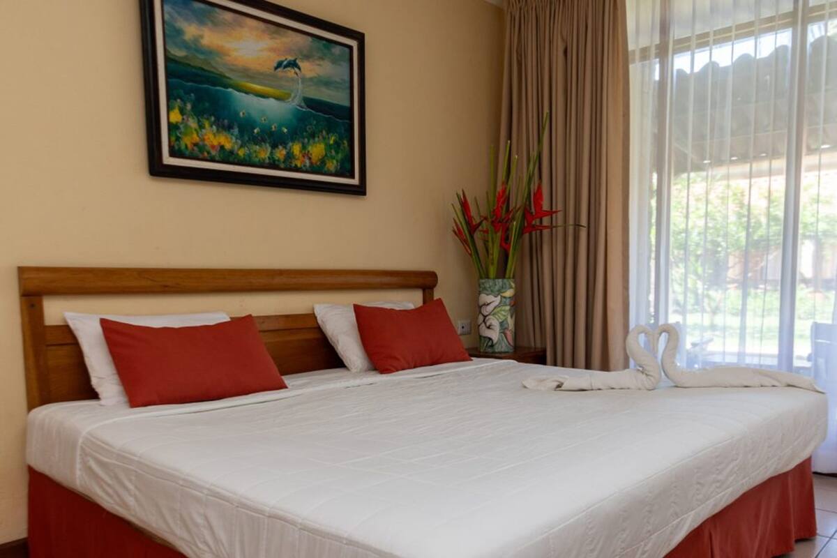 A neatly made bed with red and white pillows, a painting above it, and a vase with red flowers on the nightstand.