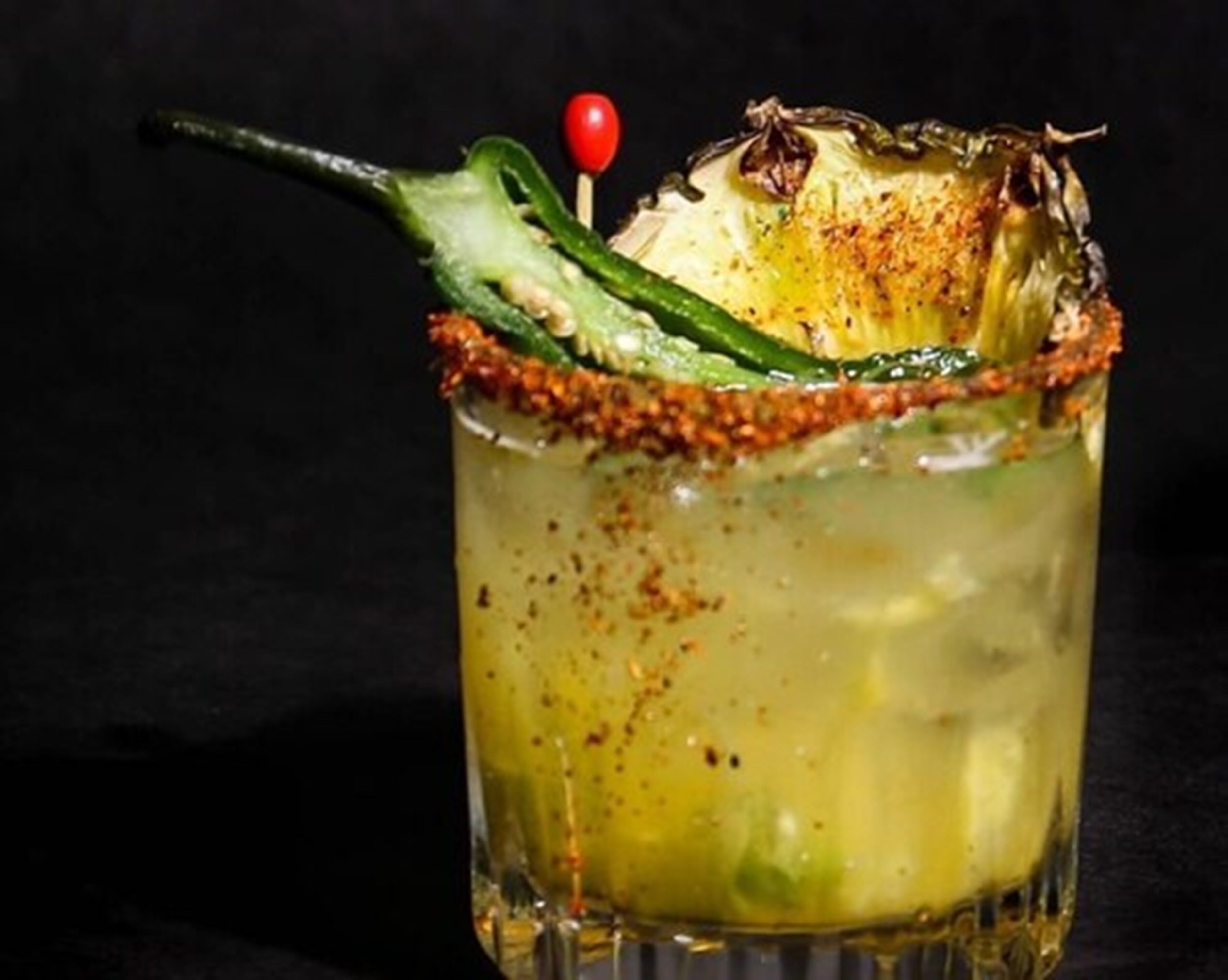 Cóctel con un chile verde y una cereza roja como adorno, servido en un vaso con borde salado.