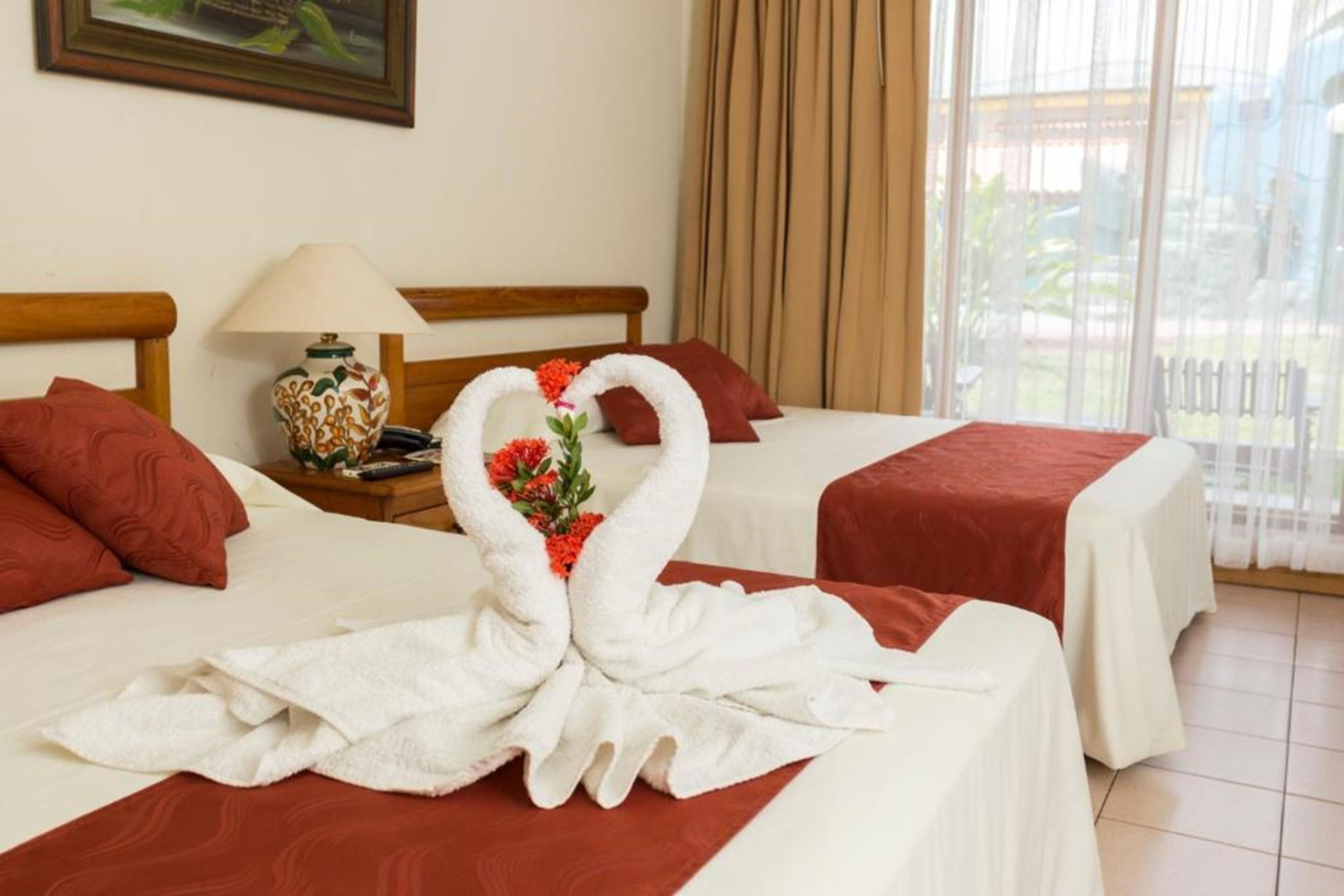 Habitación de hotel con dos camas, cada una con una almohada roja y una colcha blanca. Hay una mesita de noche con una lámpara y un jarrón decorativo.
