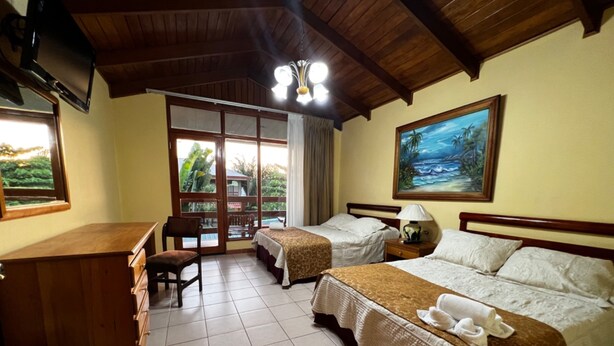 A hotel room with two beds, a desk, and a chair. There is a large window with a view of greenery outside. - Hotel Amapola Resort (Jacó)