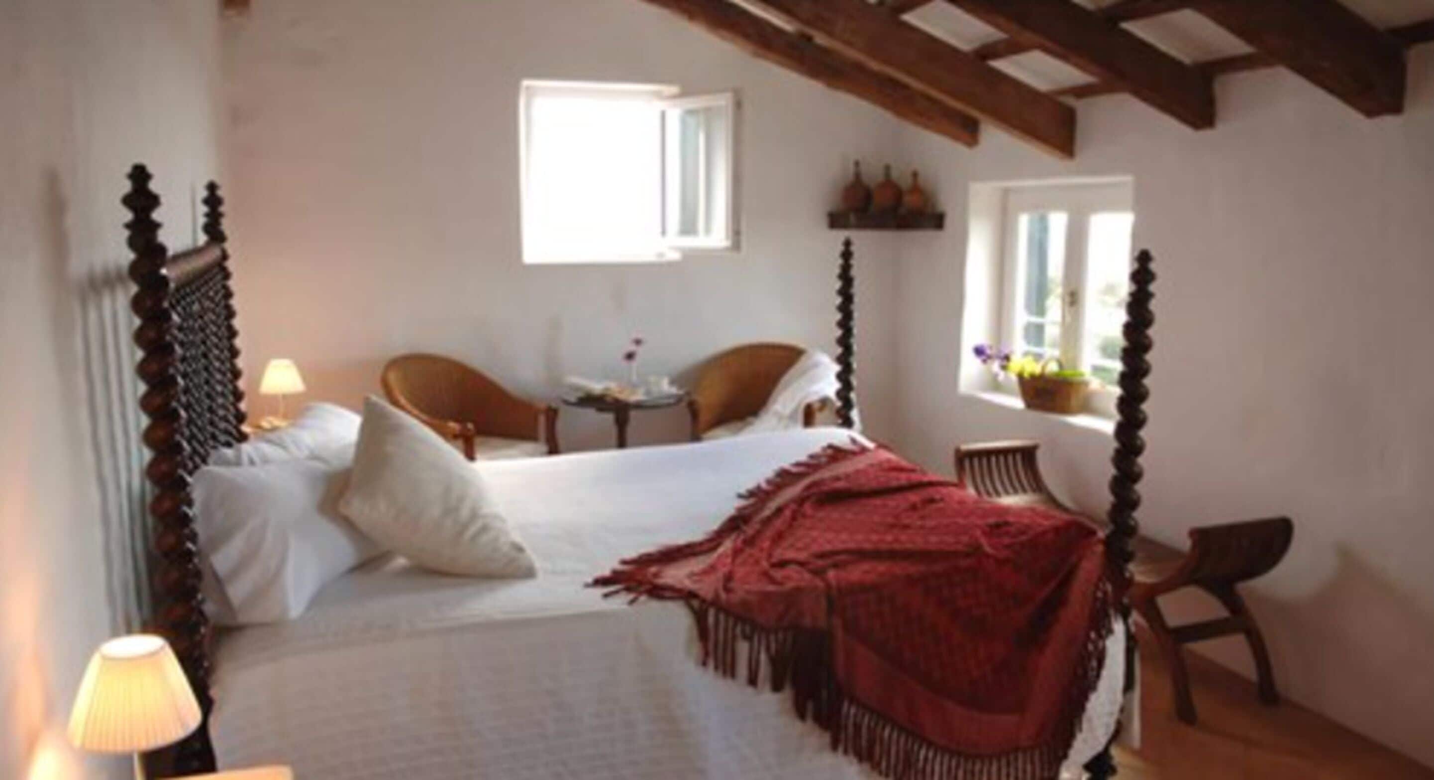 A bedroom with a four-poster bed, a small table with a chair, and a window with a view of the outside.