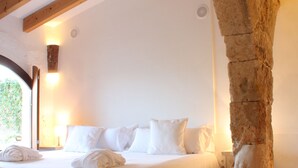A bedroom with a bed, pillows, a wooden bench, a stone pillar, and a round rug. -  Alcaufar Vell Hotel Rural & Restaurant (Sant Lluis)