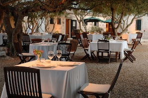 An outdoor dining area with tables set for two, surrounded by trees and a building in the background. -  Alcaufar Vell Hotel Rural & Restaurant (Sant Lluis)