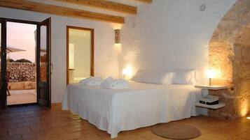 A bedroom with a stone wall, wooden beams, a bed with white linens, and a small bedside table with a lamp.