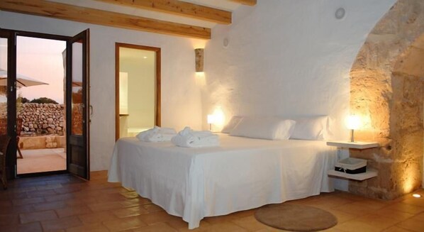 Un dormitorio con pared de piedra, vigas de madera, una cama con ropa blanca y una mesita de noche con lámpara.