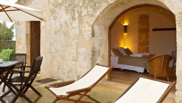 A cozy outdoor seating area with two lounge chairs and a table, an archway leading to a bedroom with a bed and a nightstand, and a wicker chair in the background. -  Alcaufar Vell Hotel Rural & Restaurant (Sant Lluis)