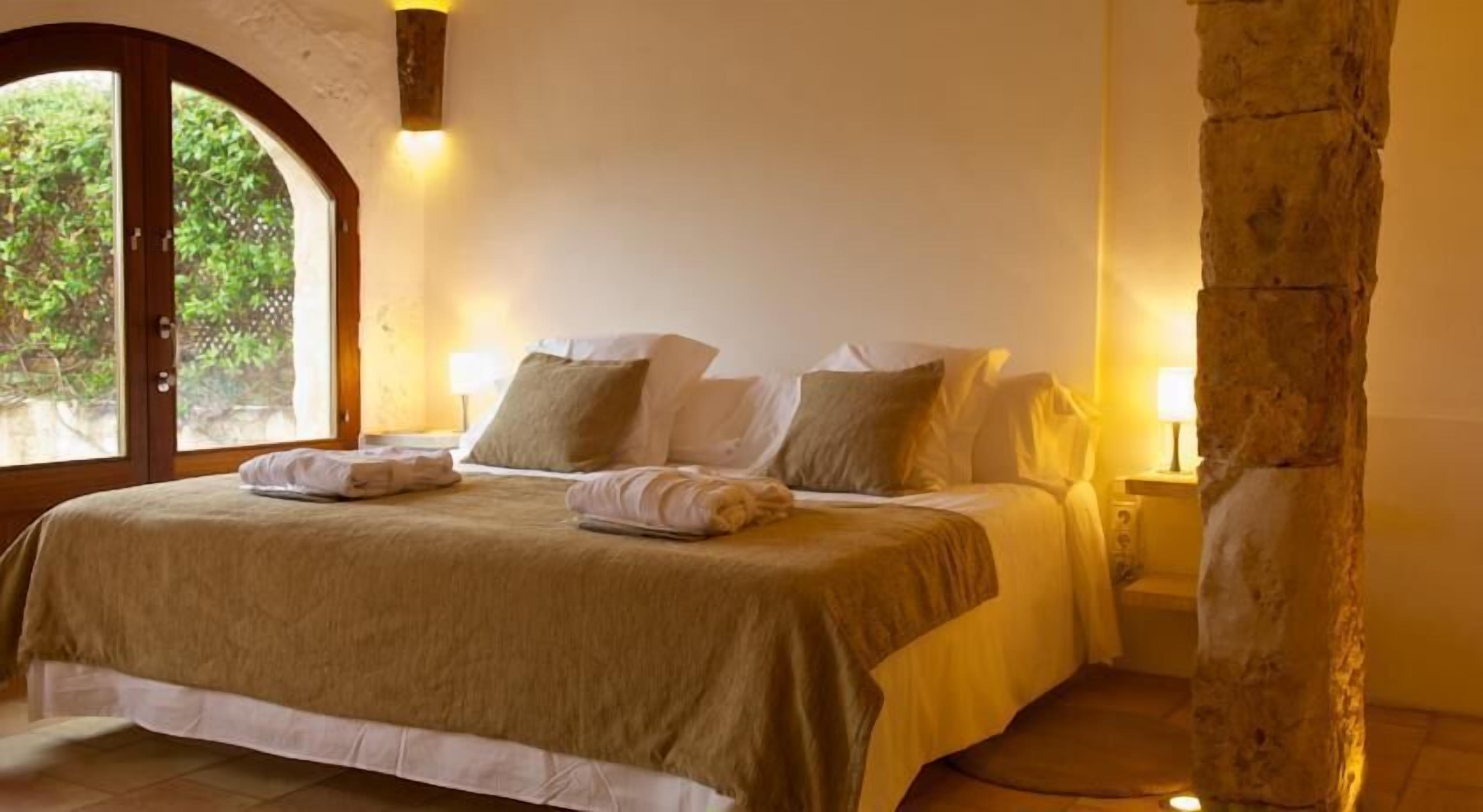 A bedroom with a large bed, two bedside lamps, a stone pillar, and a view through a window.