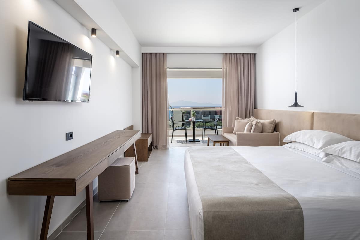 A modern hotel room with a large bed, a desk, a TV mounted on the wall, and a balcony with a table and chairs.