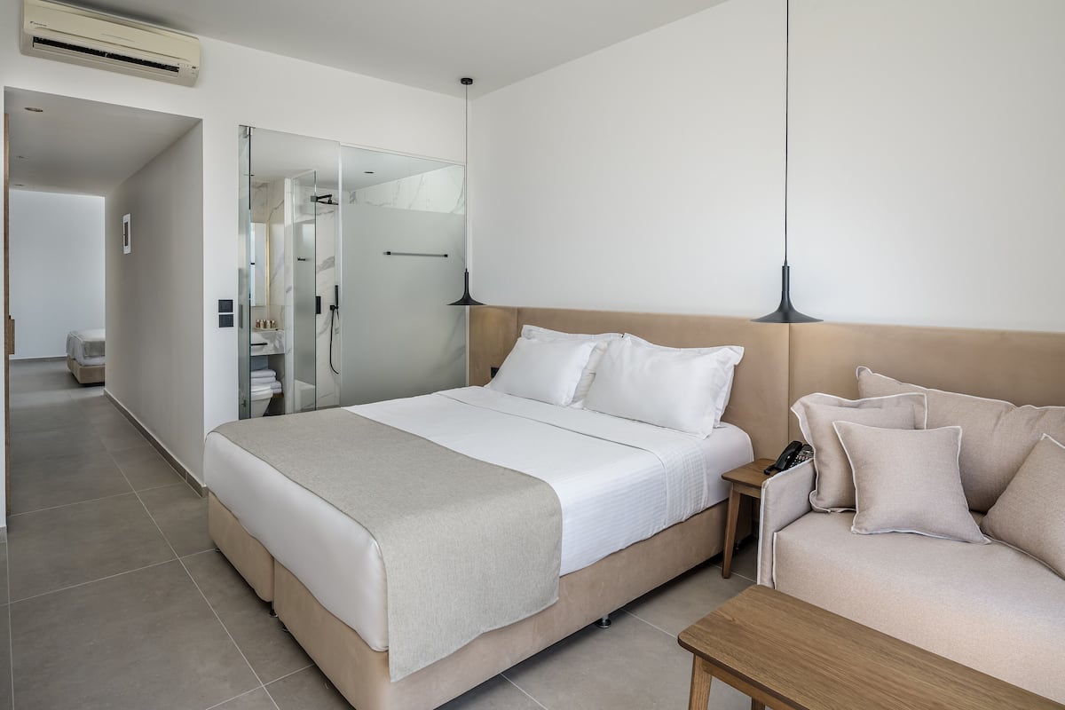 A modern hotel room with a large bed, a bedside table, a nightstand, a sofa, and a glass shower enclosure.
