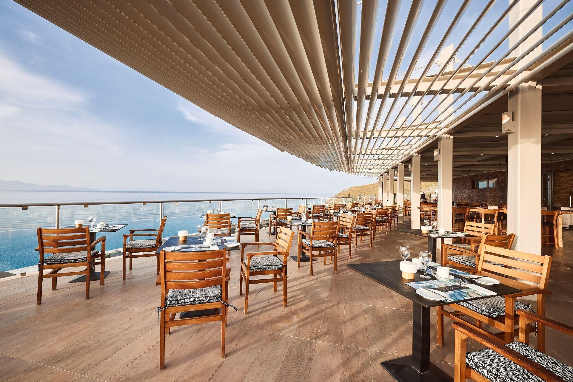 A restaurant with outdoor seating overlooking a body of water, featuring wooden tables and chairs, and a pergola structure.
