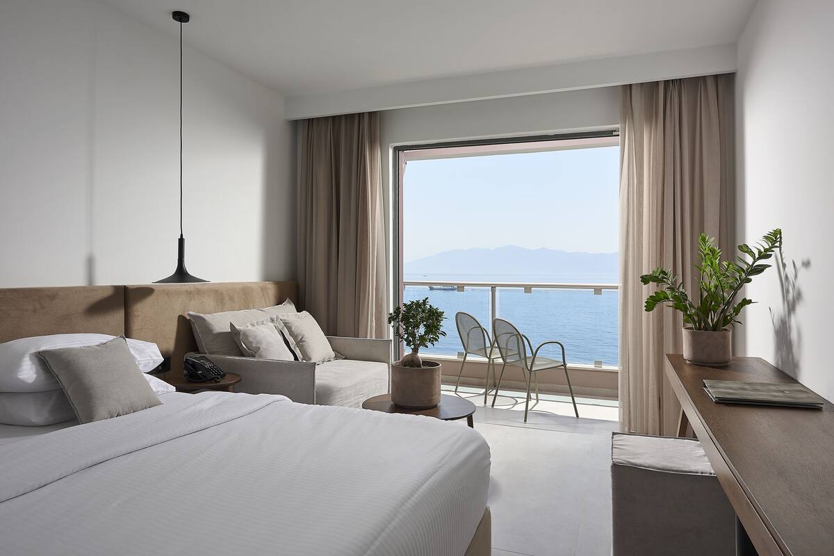 A hotel room with two beds, a desk, a chair, a potted plant, and a view of the sea.