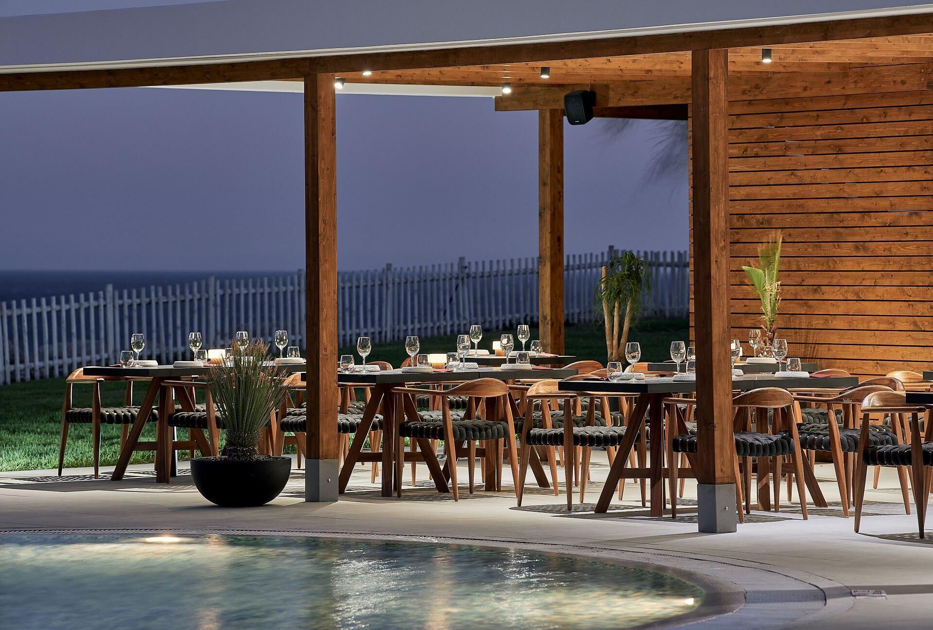 An outdoor dining area with wooden tables and chairs, a pool, and a view of the ocean.