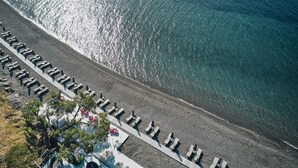 An aerial view of a beach with rows of sun loungers and umbrellas, a rocky shoreline, and clear blue water.
