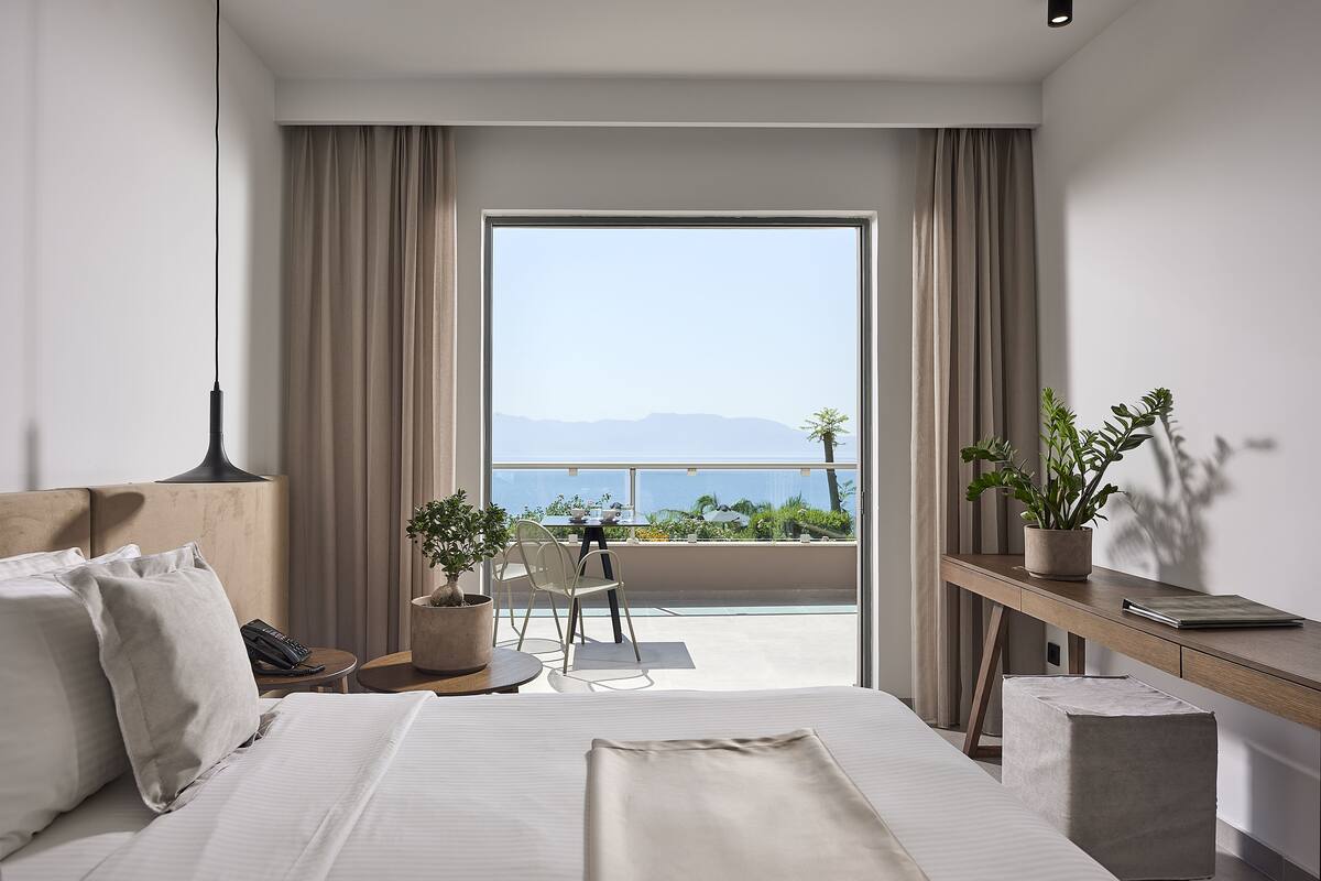 A modern hotel room with a large bed, a desk with a potted plant, a chair, and a view of the sea through a large window.