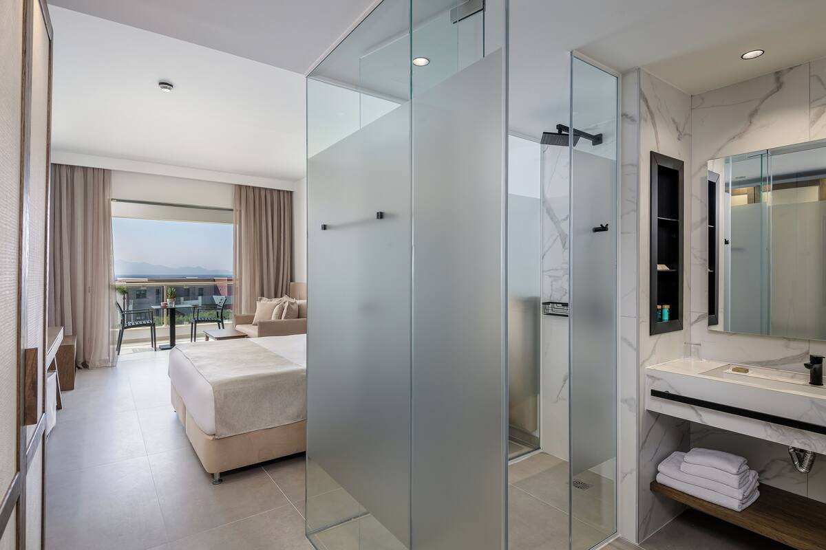 A modern hotel room with a balcony, a glass-enclosed shower, a marble bathroom, and a bed with a view.