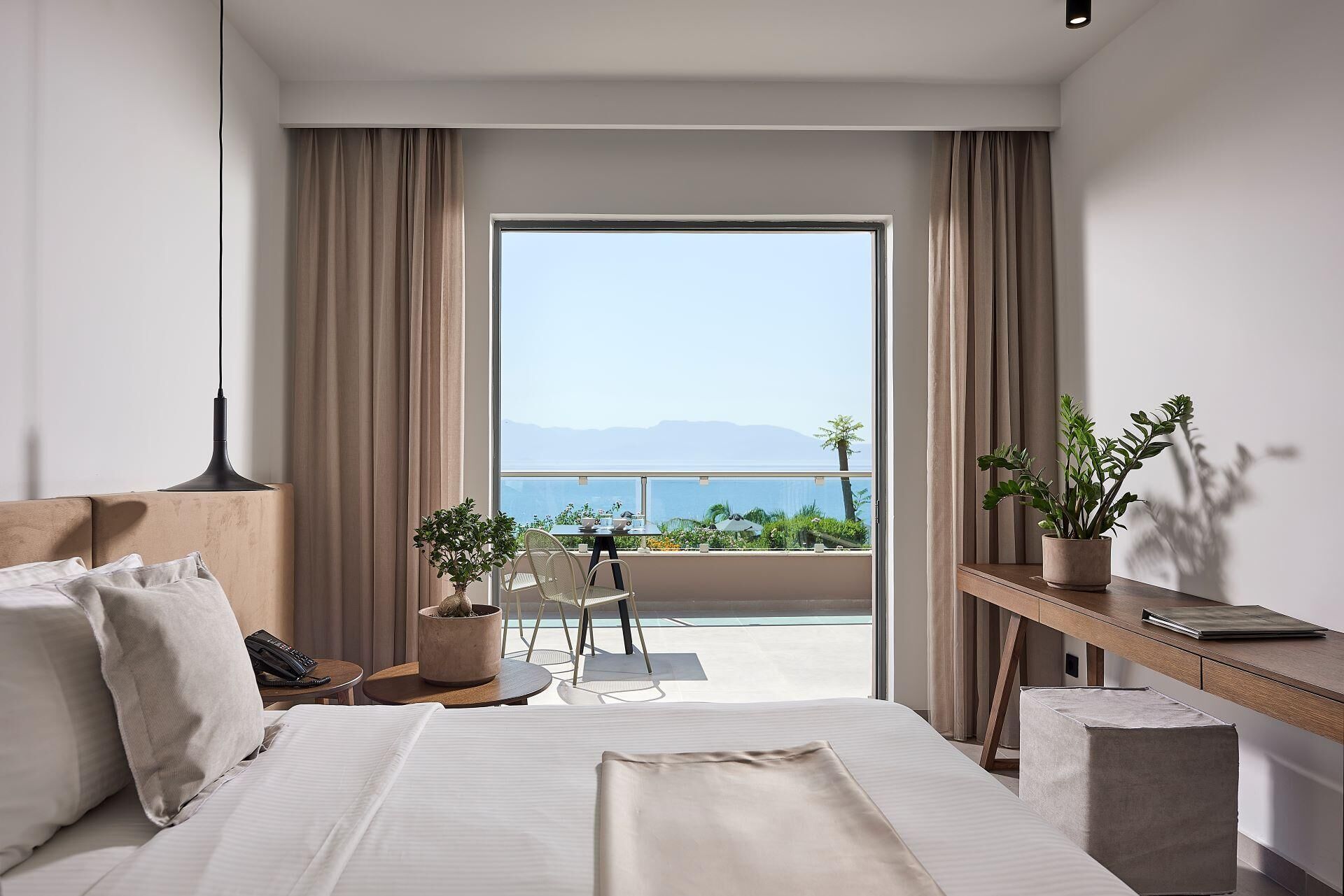 A modern hotel room with a large bed, a desk, a chair, and a balcony with a view of the sea and mountains.