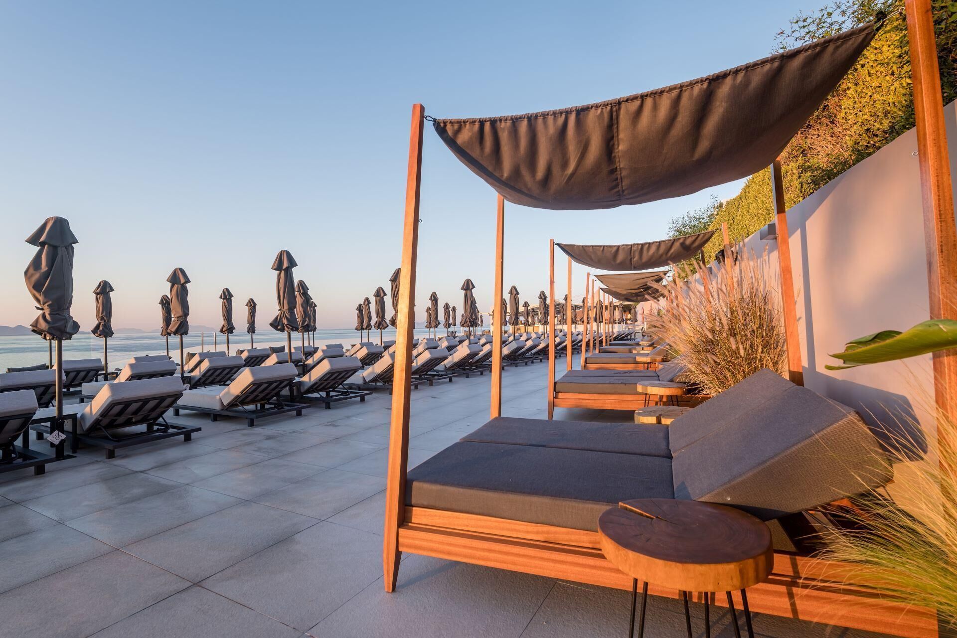 A beachside lounge area with rows of sun loungers and umbrellas, a wooden pergola structure, and a view of the sea.