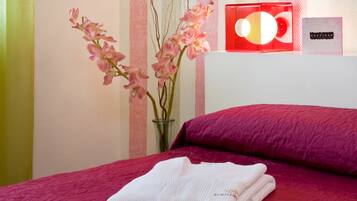 Una cama bien hecha con una colcha roja, una toalla blanca doblada, un jarrón con flores rosas y una pared a rayas rojas y blancas.