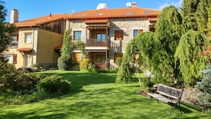 A two-story stone house with a red-tiled roof, a balcony, and a well-maintained garden with a bench. - El Milano Real (Hoyos del Espino)