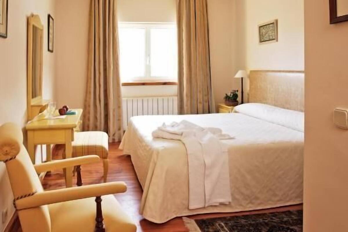 Una habitación de hotel con cama, una silla, un escritorio, una lámpara y una ventana con cortinas.